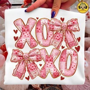 Könnte beinhalten: Weißes T-Shirt mit einem rosa-goldenen Design. Das Design zeigt die Buchstaben "XOXO" im Patchwork-Stil mit Herz- und Schleifenakzenten. Die Schleifen und Buchstaben sind mit goldenem Glitzer umrandet, mit kleinen roten Glitzerherzen.