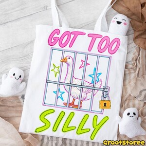 K&ouml;nnte beinhalten: Wei&szlig;e Tragetasche mit einem skurrilen Design, das eine Gans hinter Gittern zeigt, mit dem Text "GOT TOO SILLY" in Pink und Gr&uuml;n. Das Design umfasst Sterne und ein Vorh&auml;ngeschloss. Zwei kleine Geister-Pl&uuml;schtiere sind auf beiden Seiten.