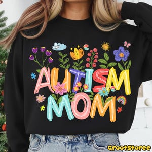 Puede incluir: Sudadera negra con la frase "AUTISM MOM" en letras coloridas y caprichosas. El texto est&aacute; rodeado de una variedad de flores, un arco&iacute;ris y otros elementos decorativos. El dise&ntilde;o est&aacute; impreso en la parte delantera de la sudadera.