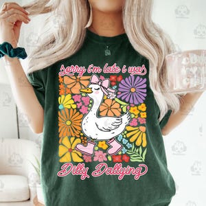 Op de afbeelding: Groen T-shirt met een witte gans die roze laarzen en een roze strik draagt. De gans is omringd door kleurrijke bloemen. De tekst "Sorry I'm late I was dilly dallying" staat op het T-shirt gedrukt.