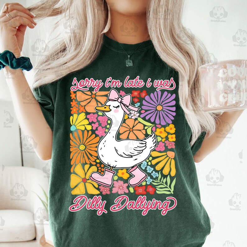 Op de afbeelding: Groen T-shirt met een witte gans die roze laarzen en een roze strik draagt. De gans is omringd door kleurrijke bloemen. De tekst "Sorry I'm late I was dilly dallying" staat op het T-shirt gedrukt.
