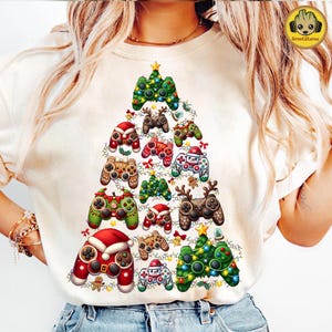 May include: A cream-coloured t-shirt displays a Christmas tree design composed of colourful video game controllers. The controllers are adorned with festive elements such as Santa hats, reindeer antlers, and fairy lights, creating a unique holiday theme.