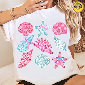 May include: White t-shirt featuring a collection of colorful seashell illustrations. The design includes pink and blue seashells, starfish with blue bows, and various shell shapes. The artwork has a hand-drawn, whimsical style.