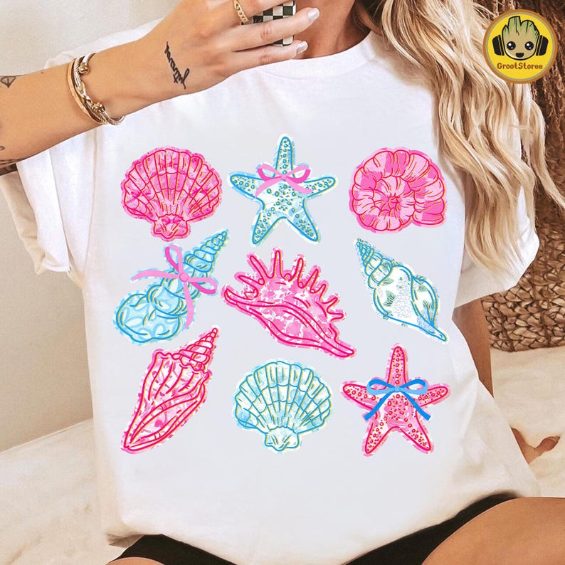 May include: White t-shirt featuring a collection of colorful seashell illustrations. The design includes pink and blue seashells, starfish with blue bows, and various shell shapes. The artwork has a hand-drawn, whimsical style.