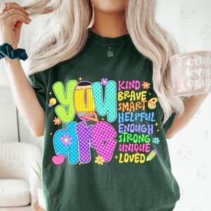 Op de afbeelding: Donkergroen t-shirt met de woorden "YOU ARE" in kleurrijke, geruite letters. Het shirt heeft ook de woorden "KIND, BRAVE, SMART, HELPFUL, ENOUGH, STRONG, UNIQUE, LOVED" met bloemen- en potloodafbeeldingen.