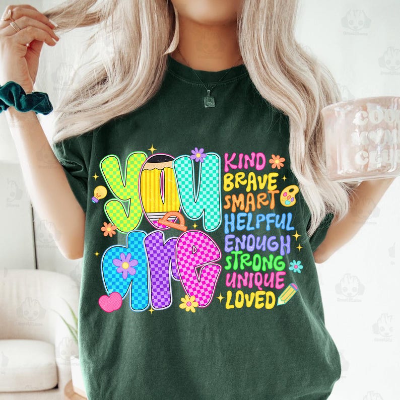 Op de afbeelding: Donkergroen t-shirt met de woorden "YOU ARE" in kleurrijke, geruite letters. Het shirt heeft ook de woorden "KIND, BRAVE, SMART, HELPFUL, ENOUGH, STRONG, UNIQUE, LOVED" met bloemen- en potloodafbeeldingen.