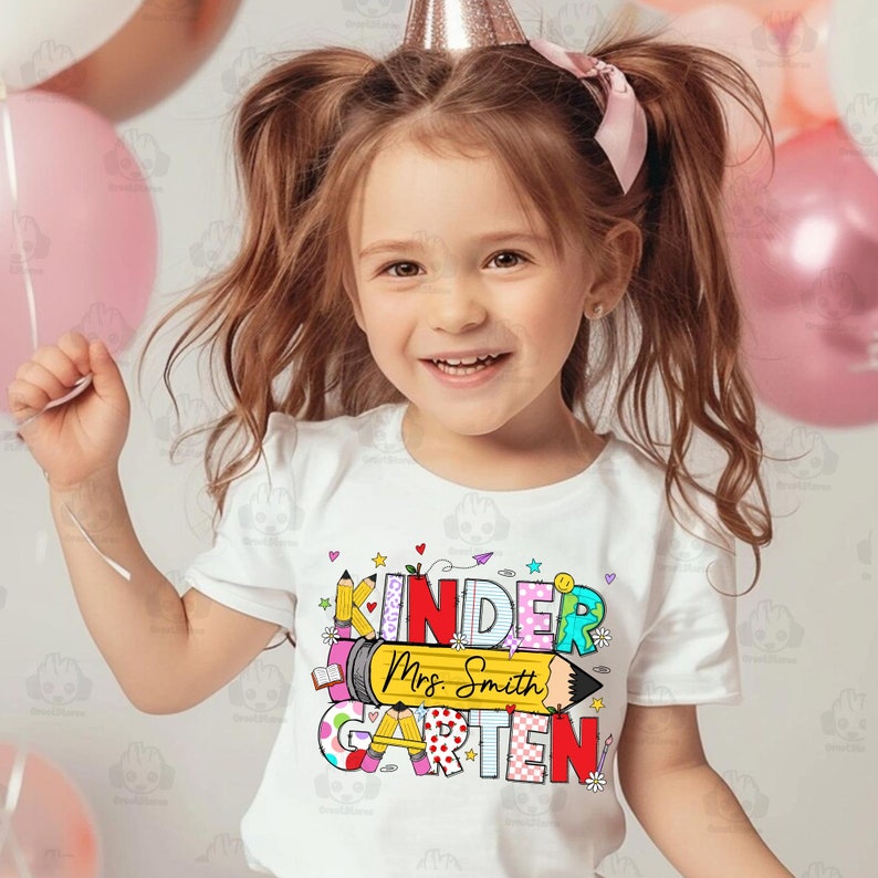 May include: A white t-shirt with a colourful graphic that says "Kinder" and "Garten" with a pencil design and the text "Mrs. Smith".