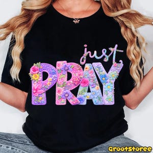 May include: Black t-shirt with the words "just PRAY" in a floral design. The word "PRAY" is in large, colorful letters with a knitted texture and filled with flowers. The word "just" is in a cursive font above "PRAY".
