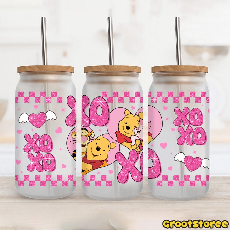 May include: Three glass tumblers with wooden lids and metal straws. Each tumbler features a pink glitter "XO XO" design with hearts and Winnie the Pooh characters. The tumblers have a clear glass body and a pink checkered pattern.