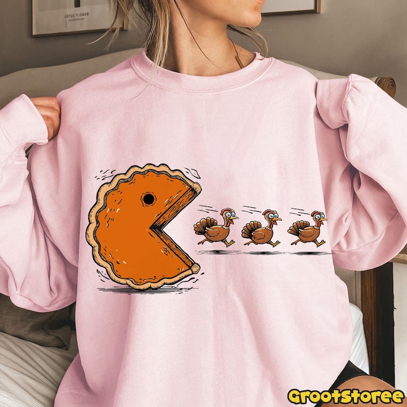 May include: A light pink sweatshirt featuring a cartoon design of a pie slice chasing three turkeys, reminiscent of the Pac-Man game. The text "GrootStoree" is printed at the bottom right.