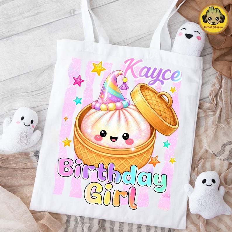 May include: A white tote bag featuring a graphic of a cute, smiling steamed bun in a bamboo steamer basket, wearing a colourful party hat. The text reads Kayce Birthday Girl in pastel colours, surrounded by small stars and pink vertical stripes.