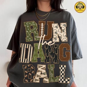Könnte beinhalten: Ein anthrazitfarbenes T-Shirt mit dem Text RUN THE DANG BALL in großen Blockbuchstaben. Jeder Buchstabe hat ein einzigartiges Muster, darunter Football-Spielzüge, Helme, Streifen, Torpfosten, Footballs, Wimpel und Schaumstofffinger in Braun und Grün.
