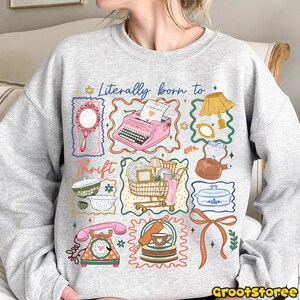 May include: A light gray sweatshirt with a colorful graphic design. The design includes a pink typewriter, a shopping cart, a telephone, and other vintage-style illustrations. Text on the shirt reads "Literally born to Thrift."