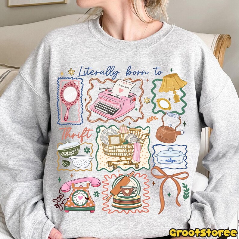 May include: A light gray sweatshirt with a colorful graphic design. The design includes a pink typewriter, a shopping cart, a telephone, and other vintage-style illustrations. Text on the shirt reads "Literally born to Thrift."