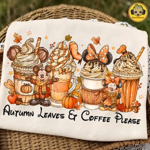 May include: A cream-colored t-shirt featuring a graphic of four iced coffee drinks decorated with autumn elements and Disney characters. The text "AUTUMN LEAVES & COFFEE PLEASE" is printed in black at the bottom. The design includes pumpkins, leaves, and acorns.