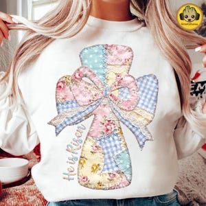 May include: White sweatshirt featuring a patchwork cross design with a bow. The cross is made of floral, polka dot, and gingham patterns in pastel colors. The text "He is Risen" is written in a script font.