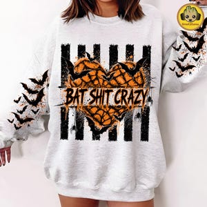 May include: A light gray crewneck sweatshirt featuring a central graphic of an orange heart made of spiderwebs, overlaid with black bats and the text BAT SHIT CRAZY. Vertical black stripes run behind the heart, and bats decorate both sleeves.
