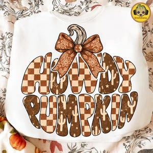Puede incluir: Una sudadera blanca de cuello redondo con un gráfico grande en forma de calabaza relleno de un patrón de cuadros marrones y beige, pequeños iconos de cactus y estrellas. El texto HOWDY PUMPKIN está integrado en el diseño, con un lazo marrón.