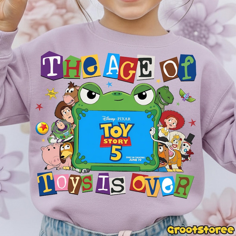 May include: A light purple sweatshirt featuring a graphic print of Toy Story characters surrounding a green frog-shaped frame. The text reads THE AGE OF TOY STORY 5 ONLY IN THEATERS JUNE 19 TOYS IS OVER in colorful, blocky, ransom-note style letters.