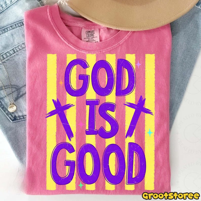 Puede incluir: Una camiseta rosa con la frase "GOD IS GOOD" en morado, sobre rayas verticales amarillas. El dise&ntilde;o incluye cruces moradas y detalles de estrellas. La camiseta es de la marca Comfort Colors.
