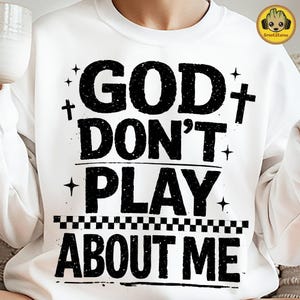 May include: White sweatshirt with the text "GOD DON'T PLAY ABOUT ME" in distressed black lettering. The design includes crosses, stars, and a checkered pattern. A casual, faith-based apparel item.