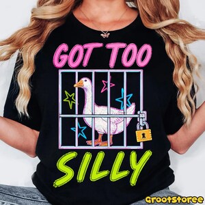K&ouml;nnte beinhalten: Schwarzes T-Shirt mit einer Grafik einer wei&szlig;en Gans in einem K&auml;fig, mit dem Text "GOT TOO SILLY" in Pink und Gr&uuml;n. Das Design beinhaltet Sterne und ein Vorh&auml;ngeschloss. Ein Freizeitkleidungsst&uuml;ck.