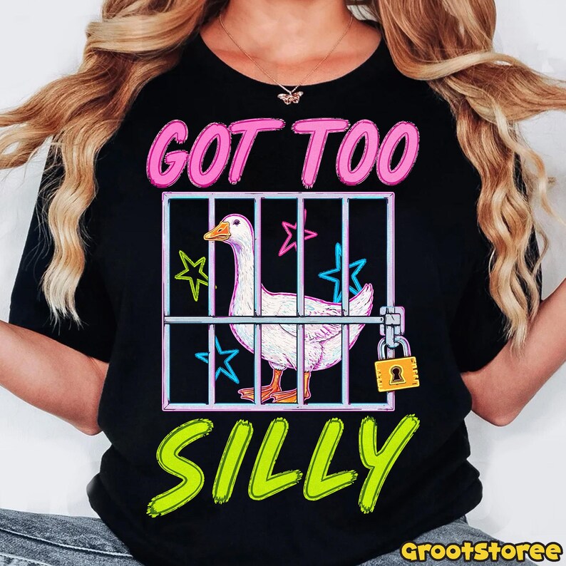 K&ouml;nnte beinhalten: Schwarzes T-Shirt mit einer Grafik einer wei&szlig;en Gans in einem K&auml;fig, mit dem Text "GOT TOO SILLY" in Pink und Gr&uuml;n. Das Design beinhaltet Sterne und ein Vorh&auml;ngeschloss. Ein Freizeitkleidungsst&uuml;ck.