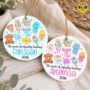 May include: Two round ceramic ornaments featuring a light blue or pink striped background with various colourful cartoon characters including a highland cow, jellyfish, and popcorn. The text reads The year of squishy hunting, Christian or Artemisia, 2026.