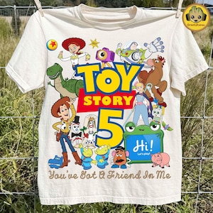 Puede incluir: Una camiseta de color crema con un estampado gráfico colorido de personajes de Toy Story, incluyendo a Woody, Buzz Lightyear, Jessie y Rex. El diseño incluye el texto "TOY STORY 5" en letras amarillas grandes y "You've Got A Friend In Me" debajo.