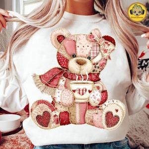 May include: White sweatshirt featuring a patchwork teddy bear design. The bear has a red scarf, a bow, and is holding a cup of coffee. The bear's patches are various shades of pink and red, with heart and floral patterns.
