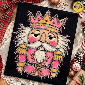 May include: A black fabric square featuring a textured, frayed-edge appliqué of a nutcracker wearing a pink and gold crown and uniform. The nutcracker has a white beard, moustache, and hair, with gold stud accents on the crown and jacket.