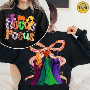 May include: Black sweatshirt with the text "Hocus Pocus" in colorful, stylized letters. The design includes images of the Sanderson sisters. The back of the sweatshirt features an illustration of the three sisters with a large bow.
