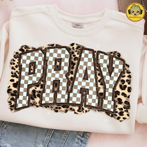 May include: Cream-colored sweatshirt with the word "PRAY" in a brown and light blue checkered design, outlined in brown. The letters are set against a leopard print background. The sweatshirt has a crew neck.