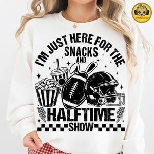 Op de afbeelding: Witte sweatshirt met een zwarte opdruk met de tekst "I'M JUST HERE FOR THE SNACKS AND HALFTIME SHOW". Het ontwerp bevat een voetbal, helm, popcorn en een beker. Een casual, comfortabele stijl.