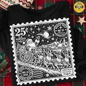 May include: A black sweatshirt featuring a white graphic print of a vintage postage stamp. The design depicts Santa Claus in a sleigh pulled by three reindeer, a snowy village, a full moon, and text including 25 cents U.S. Postage and North Pole.