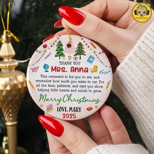 May include: A round, white Christmas ornament with a gold string, held by a person. The ornament features text that reads "THANK YOU MRS. ANNA" and "Merry Christmas." It includes illustrations of trees, books, and pencils.