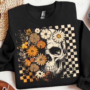 May include: Black crewneck sweatshirt featuring a skull design with floral accents. The skull is partially covered by various flowers in shades of orange, brown, and cream. A checkered pattern is on the right side of the skull.