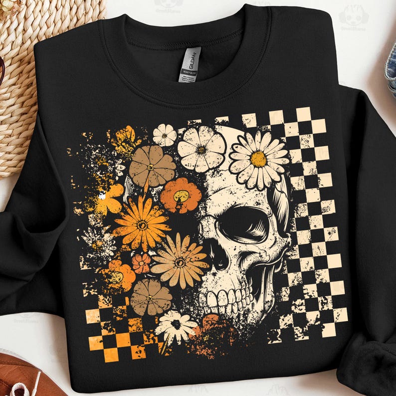 May include: Black crewneck sweatshirt featuring a skull design with floral accents. The skull is partially covered by various flowers in shades of orange, brown, and cream. A checkered pattern is on the right side of the skull.