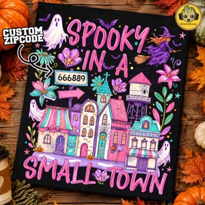 Könnte beinhalten: Ein schwarzes rechteckiges Schild mit dem Text SPOOKY IN A SMALL TOWN in rosa Buchstaben. Das Design zeigt einen weißen Geist, eine Hexe auf einem Besen, einen Wasserturm und bunte Gebäude mit Spinnweben, Blumen sowie ein Schild mit der Aufschrift 666889.