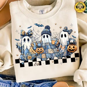Puede incluir: Sudadera color crema con un diseño de Halloween. El diseño muestra tres personajes fantasma con trajes de patchwork, sosteniendo calabazas y bebidas. Murciélagos, estrellas y telarañas completan el diseño. Un patrón de tablero de ajedrez en blanco y negro en la parte inferior.