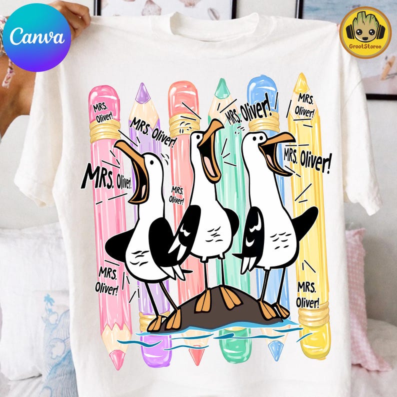 May include: A white t-shirt featuring a graphic of three seagulls standing on a rock, surrounded by five colorful pencils in pink, purple, coral, teal, and yellow. The text "MRS. OLIVER!" is printed multiple times around the birds and pencils.