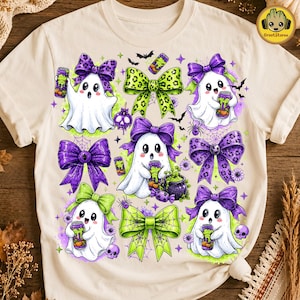 May include: A cream-colored t-shirt featuring a graphic print of four white ghosts, each holding a green energy drink can. The design includes purple and green leopard-print bows, small black bats, spiderwebs, a cauldron, and skulls.