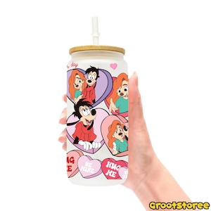 May include: A white glass can-shaped tumbler with a bamboo lid and a white straw. The tumbler features cartoon characters in heart-shaped designs and candy hearts with text. The tumbler is held by a hand.