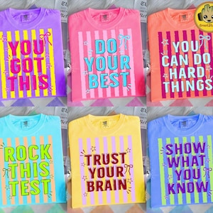 Peut inclure: Plusieurs t-shirts colorés avec des messages motivants. Chaque t-shirt présente une couleur différente et un message unique en caractères gras et ludiques. Les phrases incluent "YOU GOT THIS", "DO YOUR BEST" et "YOU CAN DO HARD THINGS".