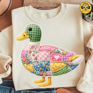 May include: Cream-colored sweatshirt featuring a patchwork duck design. The duck has a green checkered head, a yellow beak and feet, and a body composed of floral, gingham, and polka dot fabric patches. A heart is embroidered on the duck's body.