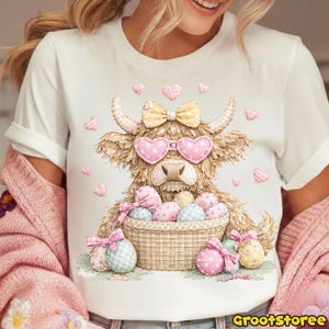 Puede incluir: Camiseta blanca con una vaca de las Tierras Altas de dibujos animados que lleva gafas de sol en forma de coraz&oacute;n y un lazo. La vaca sostiene una cesta de huevos de Pascua. Corazones rosas y huevos con lazos decoran el dise&ntilde;o. El texto "GrootStoree" est&aacute; en la esquina inferior derecha.