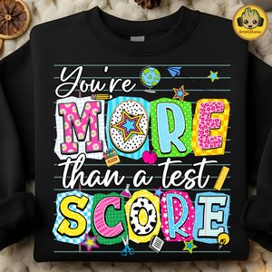 May include: Black sweatshirt with the text "You're MORE than a test SCORE" in colorful, playful letters. The design includes school-themed graphics like a globe, apple, and stars, perfect for teachers or students.
