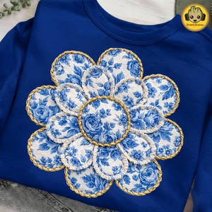 May include: A royal blue sweatshirt features a large, floral design. The flower is composed of white fabric patches with blue floral patterns, each outlined with gold rope trim. The design is centered on the chest.