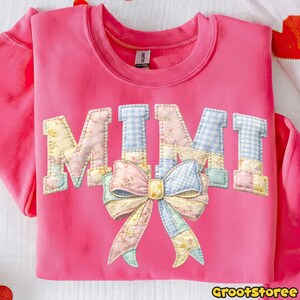 Puede incluir: Una sudadera rosa con la palabra "MIMI" en letras de patchwork. Las letras est&aacute;n decoradas con patrones florales y de cuadros, y un gran lazo est&aacute; centrado debajo. La sudadera es de cuello redondo.