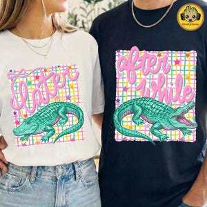 May include: White and black t-shirts featuring a teal alligator graphic with the phrases "Later" and "After a While" in pink script. The alligator is set against a colorful plaid and star background.